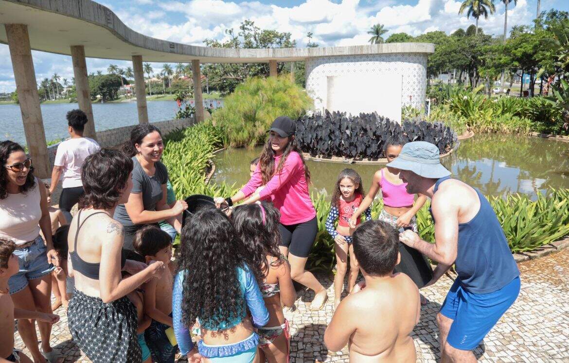 grupo de crianças e adultos brincam com baldes de água, em um dia ensolarado. Ao fundo há a estrutura e jardins da Casa do Baile