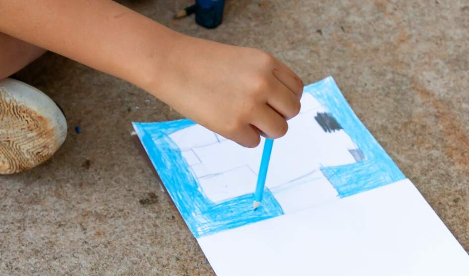 Mão de uma criança branca segura um lápis azul claro enquanto colore um papel branco