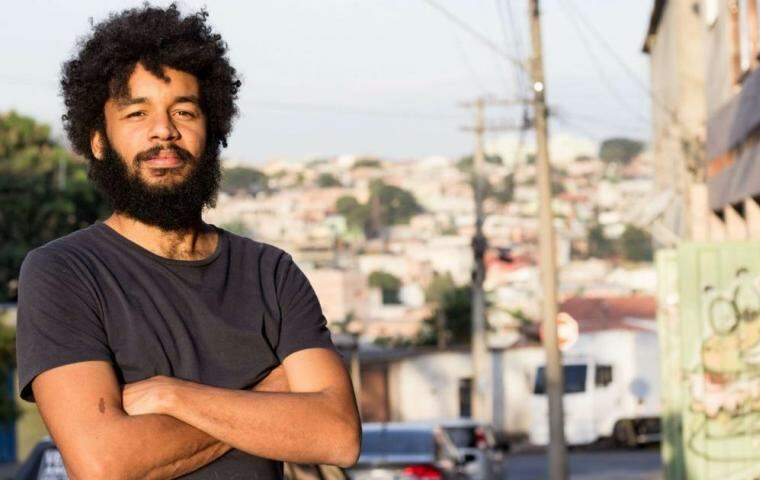 Foto do diretor do filme Marte Um, Gabriel Martins, um homem negro, barba, bigode e cabelos pretos, vestido com uma camiseta preta, de braços cruzados, posando em uma paisagem urbana desfocada.