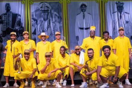 Foto estão os integrantes do bloco Babadan Banda de Rua (BH) em posição semelhante à time de futebol. Ao todo, 13 homens estão vestidos com roupas amarelas, estão sorrindo e segurando instrumentos de percussão. 
