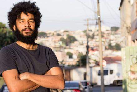 Foto do diretor do filme Marte Um, Gabriel Martins, um homem negro, barba, bigode e cabelos pretos, vestido com uma camiseta preta, de braços cruzados, posando em uma paisagem urbana desfocada.